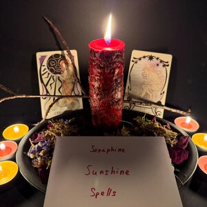May include: A red pillar candle with a textured surface is lit, surrounded by dried flowers, twigs, and small tea light candles. Two tarot cards stand behind the candle. A white card in the foreground features the handwritten text Seraphine Sunshine Spells.