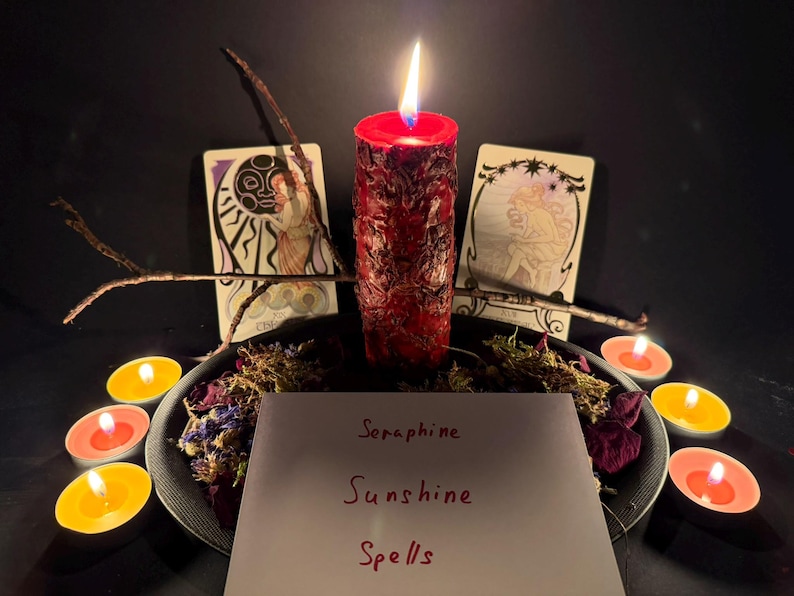 May include: A red pillar candle with a textured surface is lit, surrounded by dried flowers, twigs, and small tea light candles. Two tarot cards stand behind the candle. A white card in the foreground features the handwritten text Seraphine Sunshine Spells.
