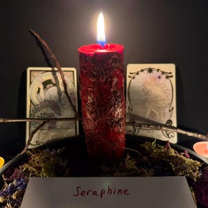Puede incluir: Una vela de pilar roja y texturizada con una llama encendida, rodeada de musgo seco, p&eacute;talos de flores y peque&ntilde;as velas de t&eacute;. Detr&aacute;s de la vela hay dos cartas del tarot y una ramita. Una tarjeta blanca en primer plano muestra el texto Seraphine.