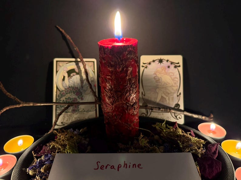 Puede incluir: Una vela de pilar roja y texturizada con una llama encendida, rodeada de musgo seco, p&eacute;talos de flores y peque&ntilde;as velas de t&eacute;. Detr&aacute;s de la vela hay dos cartas del tarot y una ramita. Una tarjeta blanca en primer plano muestra el texto Seraphine.
