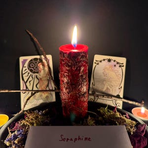 May include: A red pillar candle with a textured, uneven surface is lit, casting a warm glow. It is surrounded by dried flowers, small tea light candles, two illustrated cards featuring celestial figures, and a twig. A white card reads Seraphine.