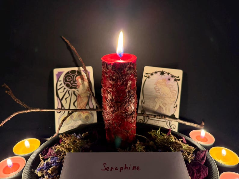 May include: A red pillar candle with a textured, uneven surface is lit, casting a warm glow. It is surrounded by dried flowers, small tea light candles, two illustrated cards featuring celestial figures, and a twig. A white card reads Seraphine.