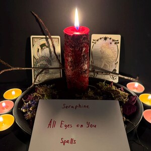 Puede incluir: Una vela de pilar roja con superficie texturizada est&aacute; encendida, rodeada de flores secas y peque&ntilde;as velas de t&eacute;. Dos cartas del tarot se encuentran detr&aacute;s de la vela. Una tarjeta blanca en primer plano muestra el texto Seraphine All Eyes on You Spells.