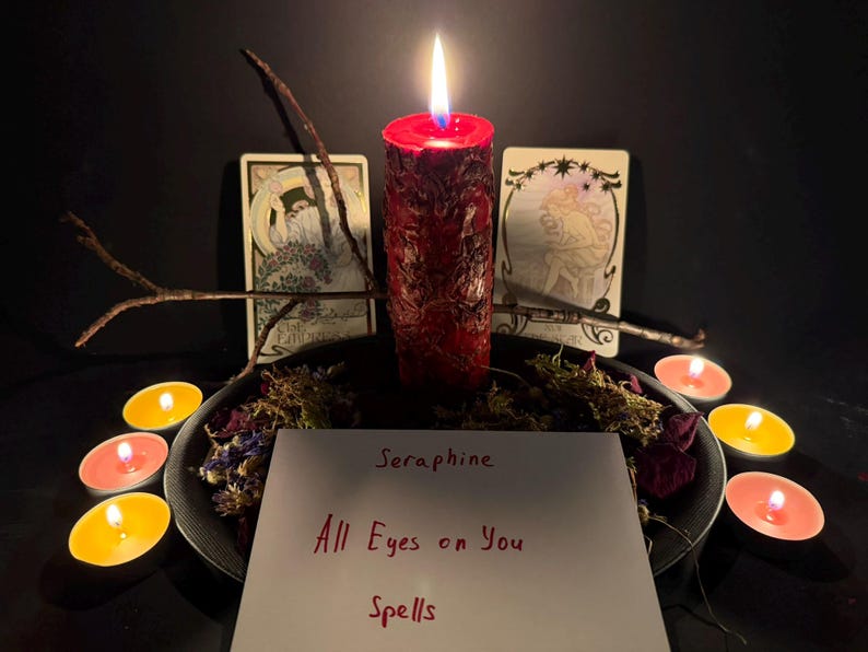 Puede incluir: Una vela de pilar roja con superficie texturizada est&aacute; encendida, rodeada de flores secas y peque&ntilde;as velas de t&eacute;. Dos cartas del tarot se encuentran detr&aacute;s de la vela. Una tarjeta blanca en primer plano muestra el texto Seraphine All Eyes on You Spells.