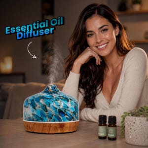 Pode incluir: Um difusor de óleos essenciais com um topo de vidro de mosaico azul e branco e uma base de madeira clara, emitindo um fino fluxo de névoa. Dois pequenos frascos de vidro âmbar com tampas pretas estão ao lado do difusor numa superfície de madeira.