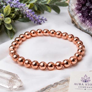 May include: A circular bracelet composed of polished, metallic copper-colored spherical beads. The bracelet is arranged on a white fabric surface alongside a clear quartz crystal, a purple amethyst geode, lavender sprigs, and a small white card.