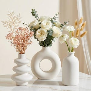 Può includere: Tre vasi in ceramica bianca di forme diverse con composizioni floreali. Il vaso a sinistra ha una silhouette ondulata e impilata con steli secchi rosa e bianchi. Il vaso centrale è circolare con un centro cavo, riempito di rose bianche ed eucalipto. Il vaso a destra è cilindrico e contiene ranuncoli bianchi e code di lepre color marrone chiaro.
