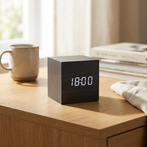 Può includere: Una sveglia digitale a forma di cubo con una finitura in venature di legno scuro. La parte anteriore presenta un luminoso display a LED bianco che mostra l'ora 18:00. L'orologio ha una struttura solida e liscia con bordi netti e un aspetto.