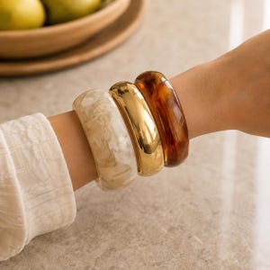 Op de afbeelding: Een set van drie dikke armbanden die samen worden gedragen. Eén armband is van crèmekleurige gemarmerde hars, de tweede is van gepolijst goudkleurig metaal en de derde is van donkerbruine hars met een schildpadpatroon en een glanzende afwerking.