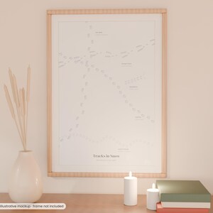 May include: A framed wall art print titled Tracks in Snow featuring a white background with various gray animal footprint patterns and labels. The print is displayed in a light wood frame alongside a white vase with dried stems and candles.