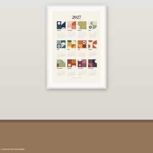 May include: A 2027 wall calendar print featuring twelve monthly grids, each topped with a unique geometric square design in shades of blue, red, yellow, and green on a cream background, displayed within a white frame on a wall.