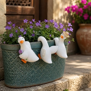 May include: A teal oval planter with a textured diamond pattern, featuring three white goose figurines with orange beaks and feet perched on the rim. One goose holds a small purple flower, and another wears a tiny tan straw hat.