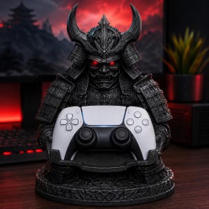 May include: A dark gray samurai warrior figurine featuring a detailed helmet with horns and glowing red eyes. The figure is designed as a stand, holding a white and black wireless video game controller in its hands on a circular stone-textured base.