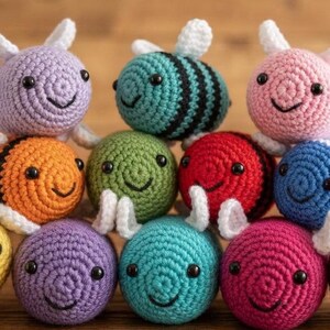 Puede incluir: Una colección de once peluches redondos con forma de abeja tejidos a ganchillo en varios colores, incluyendo amarillo, morado, naranja, verde, rojo, azul, rosa y marrón. Cada peluche tiene pequeñas alas blancas, ojos de cuentas negras y una sonrisa bordada.