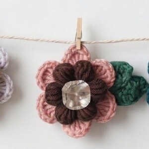 May include: Three handmade crochet flowers hanging from a string with wooden clips. The first flower has light purple petals, a mustard yellow center, and a dark brown button. The second has pink petals, a dark brown center, and a clear crystal. The third has teal petals, a rust-colored center, and a clear crystal, all with dark green leaves.
