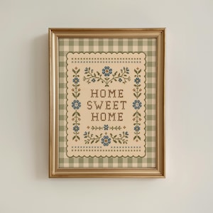 Puede incluir: Un bordado en punto de cruz enmarcado con el texto "HOME SWEET HOME" en letras marrones. El diseño incluye motivos florales azules y enredaderas verdes sobre un fondo crema con un borde festoneado, todo dentro de un marco dorado.