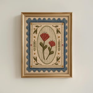 Op de afbeelding: Een ingelijst textielkunstwerk met een geborduurd ovaal middenstuk met twee rode bloemen op een lichtgroene gestreepte achtergrond. Het ontwerp is omgeven door een crèmekleurige rand met kleine bloemmotieven en een geschulpte blauwe rand.