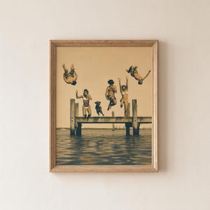 Könnte beinhalten: Ein gerahmter Kunstdruck in Sepiatönen zeigt einen Holzsteg, der in das Wasser ragt. Mehrere Personen und ein Hund sind in der Luft zu sehen, wie sie vom Steg ins Wasser springen. Die Szene ist in hellbraunen Farbtönen mit dunklen Silhouetten gehalten.