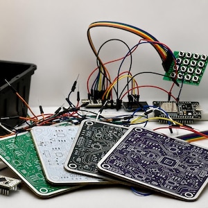 May include: A collection of four square printed circuit boards in green, white, black, and purple, featuring intricate silver circuitry patterns. Nearby are electronic components including a breadboard, a button matrix, and colorful jumper wires.