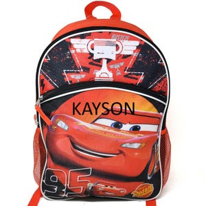May include: An orange and black backpack featuring a graphic of a red race car with the number 95 and the name KAYSON printed in black. The bag includes a top handle, a zippered front pocket, and orange mesh side pockets for storage.