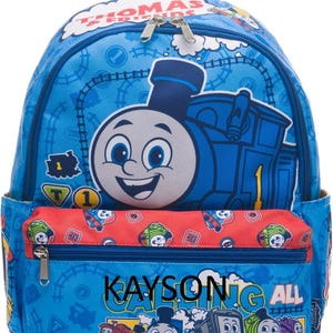 May include: A blue Thomas & Friends backpack featuring a large Thomas the Tank Engine graphic, a red front pocket with a repeating train pattern, and the text Kayson Calling All Engines. The bag includes silver zippers and a top handle.