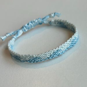 May include: A woven friendship bracelet featuring a repeating wave pattern in shades of light blue and white. The bracelet is constructed from thin thread and includes adjustable knotted ends for securing the piece around a wrist or ankle.