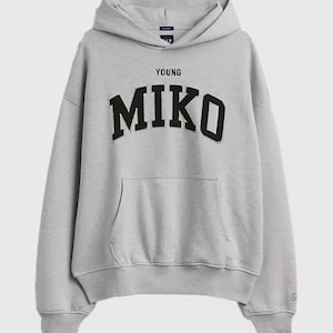 May include: A light gray long-sleeve hooded sweatshirt featuring a front kangaroo pocket and ribbed cuffs. The text YOUNG MIKO is printed in black block lettering across the chest. A small Gap brand logo is visible on the inner neck label.