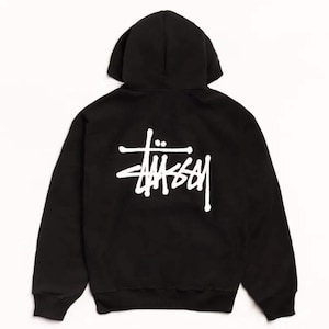 May include: A black hooded sweatshirt featuring a white Stüssy logo printed on the back. The garment includes a hood, long sleeves with ribbed cuffs, and a ribbed hem, showcasing the brand's signature script graphic centered on the upper back area.