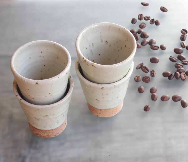 May include: Four handmade ceramic coffee cups with a speckled white glaze and a reddish-brown base. Coffee beans are scattered around the cups. The cups are stacked in pairs, showcasing their unique, rustic design.
