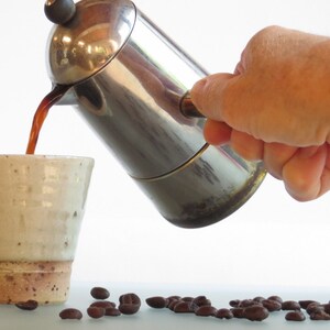 May include: A stainless steel stovetop espresso maker pouring dark coffee into a speckled ceramic mug. Coffee beans are scattered on the surface. The mug has a light beige color with a brown base.