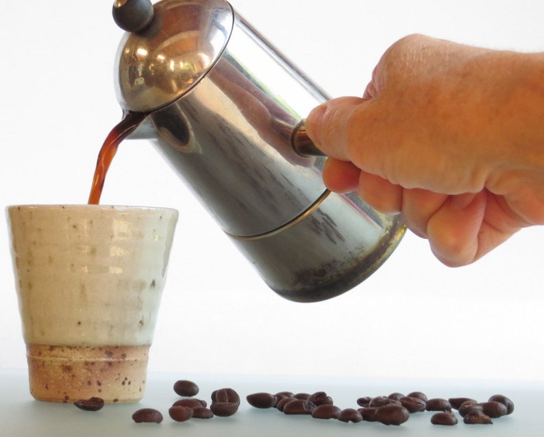 May include: A stainless steel stovetop espresso maker pouring dark coffee into a speckled ceramic mug. Coffee beans are scattered on the surface. The mug has a light beige color with a brown base.