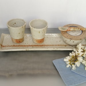 May include: A set of three ceramic cups and a small bowl with a spoon, all in a speckled white glaze with brown accents, arranged on a rectangular tray with a brown stripe. The tray is on a gray surface, and a blue cloth with white flowers is in the foreground.