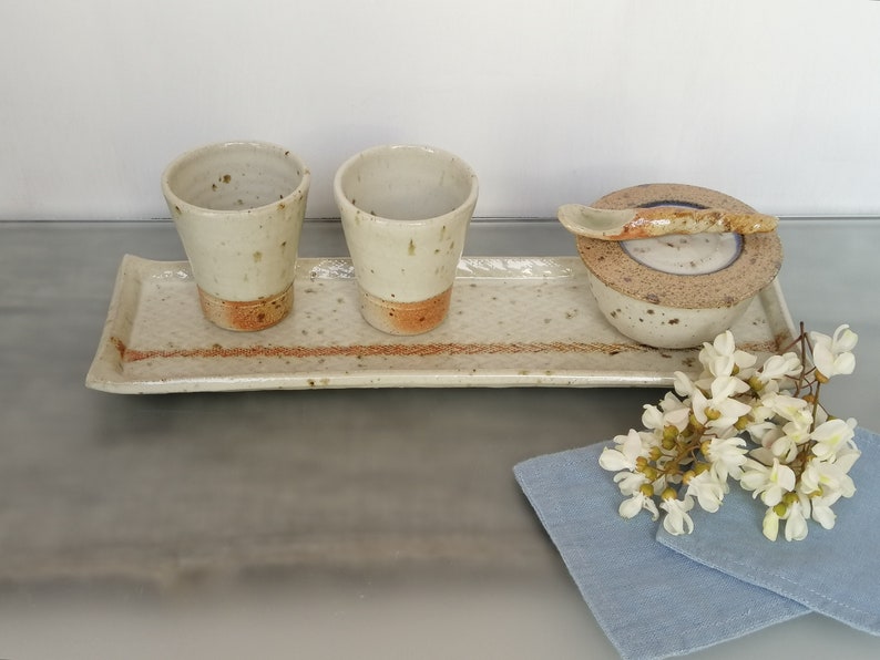 May include: A set of three ceramic cups and a small bowl with a spoon, all in a speckled white glaze with brown accents, arranged on a rectangular tray with a brown stripe. The tray is on a gray surface, and a blue cloth with white flowers is in the foreground.