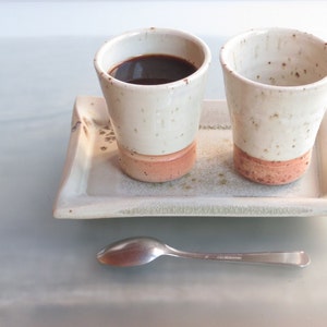 May include: Two ceramic coffee cups with a white speckled glaze and a reddish-brown base sit on a rectangular tray. One cup is filled with dark coffee. A silver spoon rests on the surface below the tray. The set is ideal for serving coffee or tea.