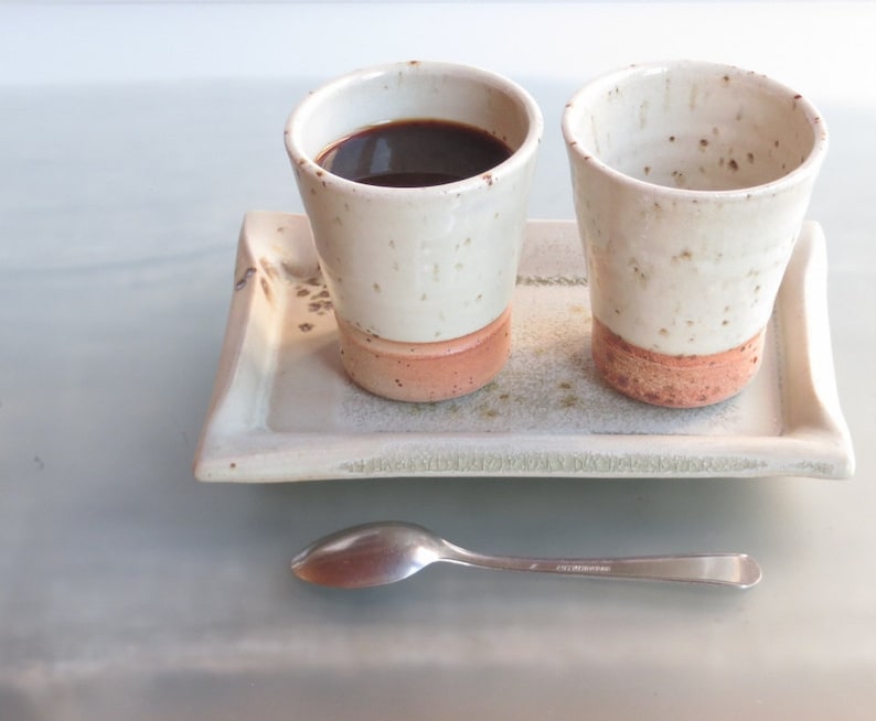 May include: Two ceramic coffee cups with a white speckled glaze and a reddish-brown base sit on a rectangular tray. One cup is filled with dark coffee. A silver spoon rests on the surface below the tray. The set is ideal for serving coffee or tea.
