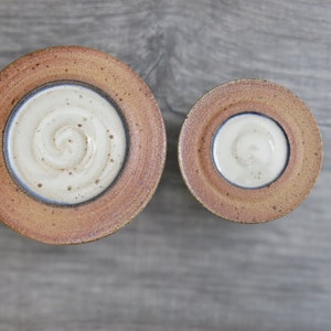 May include: Two ceramic bowls with a brown glaze and a white interior. The larger bowl has a swirl design in the center. The smaller bowl has a plain white interior.