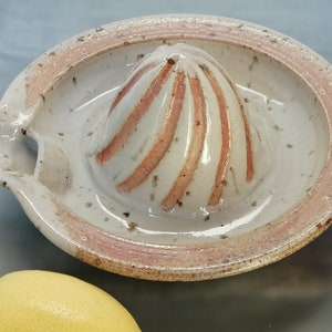 Op de afbeelding: Een witte keramische citruspers met een rood-wit gestreept ontwerp. De pers heeft een tuit om sap uit te gieten en een verhoogde rand.