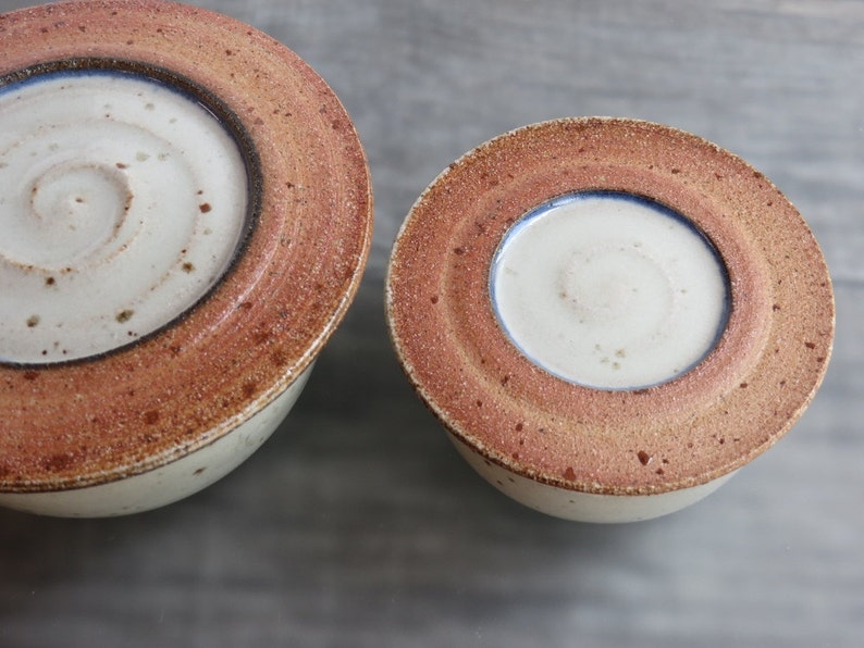 May include: Two ceramic bowls with lids. The bowls are white with a brown rim and a blue line around the inside. The lids are the same color and have a similar design.