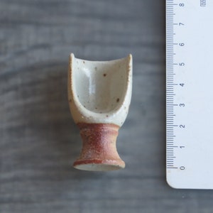 May include: A small, white ceramic cup with a brown base. The cup is designed to hold a single serving of something, like a teacup.
