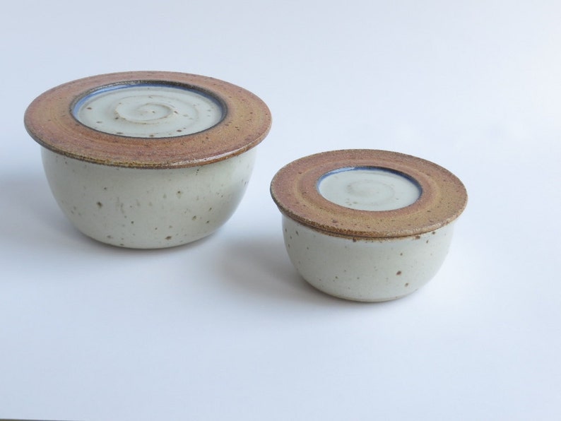 May include: Two ceramic bowls with lids. The larger bowl is white with brown speckles and a brown lid with a blue rim. The smaller bowl is white with brown speckles and a brown lid with a blue rim.