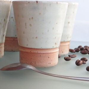 May include: Four handmade ceramic coffee cups with a speckled white glaze and a reddish-brown base. A silver spoon rests in front of the cups, with scattered coffee beans nearby. The cups have a simple, modern design.
