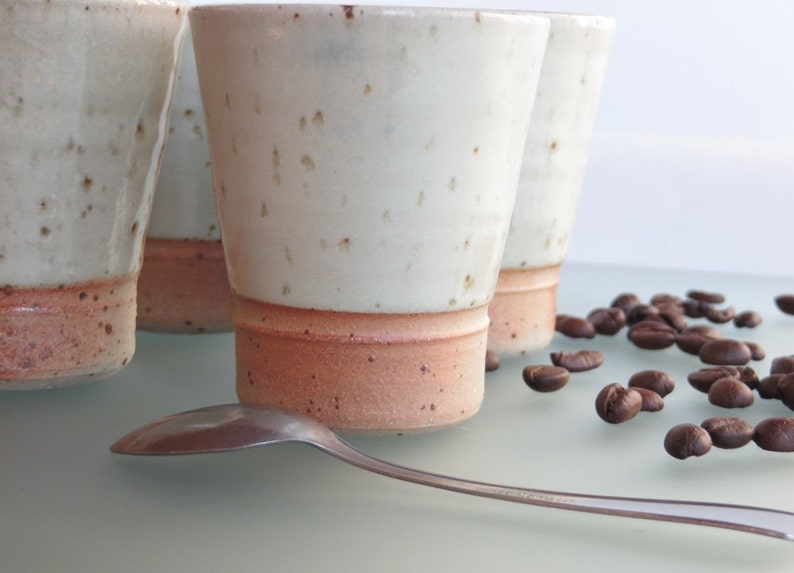 May include: Four handmade ceramic coffee cups with a speckled white glaze and a reddish-brown base. A silver spoon rests in front of the cups, with scattered coffee beans nearby. The cups have a simple, modern design.