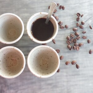 May include: Four small, speckled ceramic cups, one filled with dark coffee and a spoon. Roasted coffee beans are scattered around the cups on a light gray surface. The cups have a simple, minimalist design.