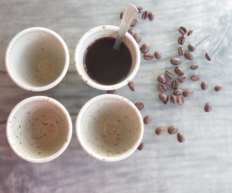 May include: Four small, speckled ceramic cups, one filled with dark coffee and a spoon. Roasted coffee beans are scattered around the cups on a light gray surface. The cups have a simple, minimalist design.