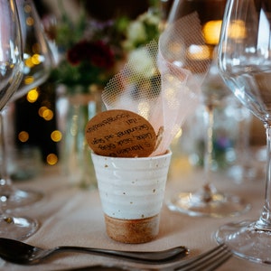 May include: A white ceramic cup with a brown base holds a decorative item with text, set on a table with glassware and a spoon. The background features blurred flowers and lights, suggesting a celebratory event.