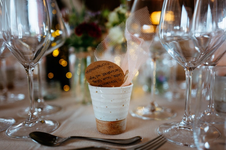 May include: A white ceramic cup with a brown base holds a decorative item with text, set on a table with glassware and a spoon. The background features blurred flowers and lights, suggesting a celebratory event.