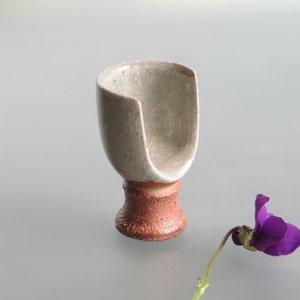 May include: A small, white ceramic cup with a unique, asymmetrical shape and a brown base. The cup is sitting on a light gray surface next to a purple flower.