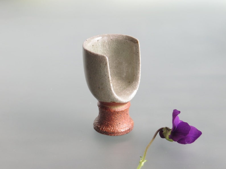 May include: A small, white ceramic cup with a unique, asymmetrical shape and a brown base. The cup is sitting on a light gray surface next to a purple flower.