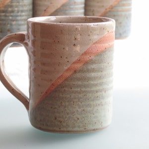 Może przedstawiać: Ręcznie robiony ceramiczny kubek z beżowym i szarym nakrapianym wykończeniem. Przekątna wstęga z koralowej glazury podkreśla powierzchnię kubka. Kubek ma zakrzywiony uchwyt i jest częścią zestawu podobnych kubk&oacute;w.