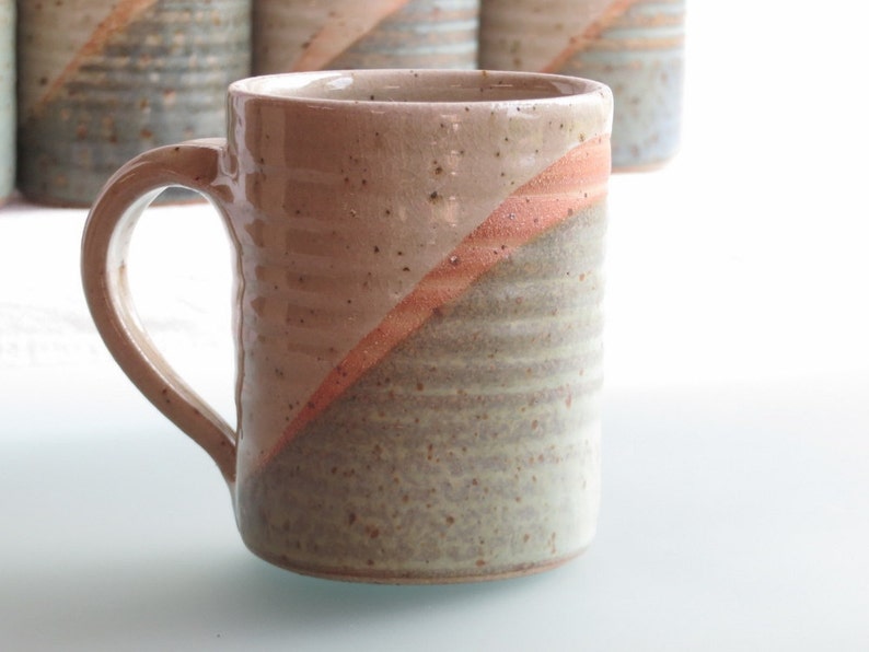 Może przedstawiać: Ręcznie robiony ceramiczny kubek z beżowym i szarym nakrapianym wykończeniem. Przekątna wstęga z koralowej glazury podkreśla powierzchnię kubka. Kubek ma zakrzywiony uchwyt i jest częścią zestawu podobnych kubk&oacute;w.